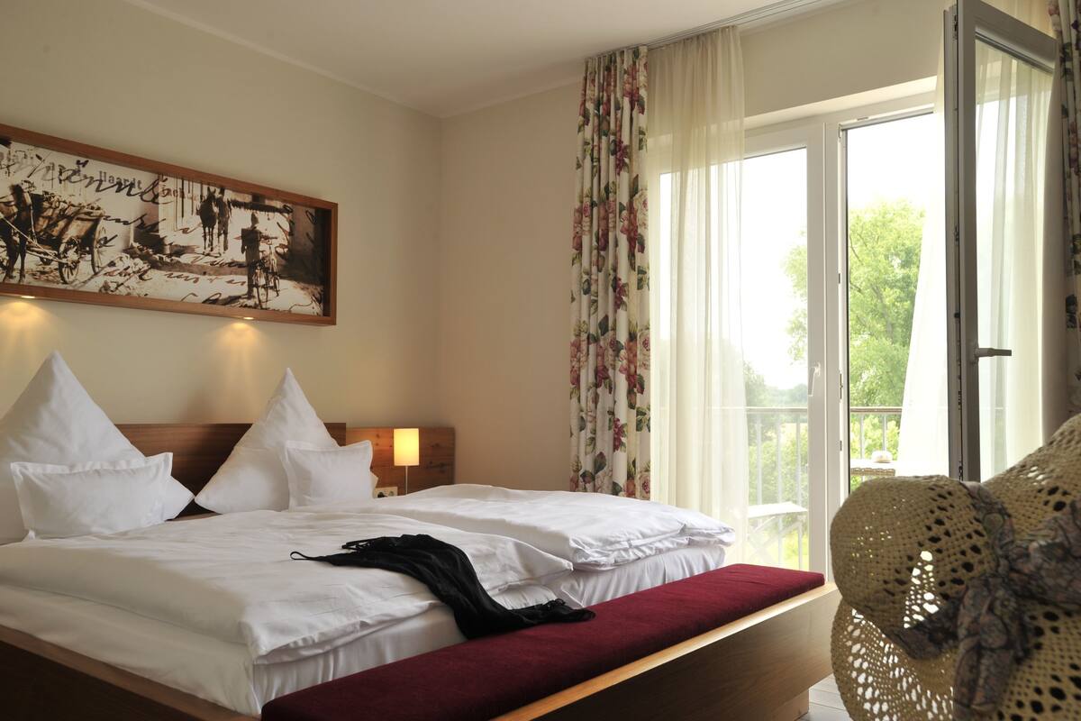 Une chambre d’hôtel avec un lit, une table de chevet, un tableau, une fenêtre avec des rideaux et une vue sur la verdure à l’extérieur.
