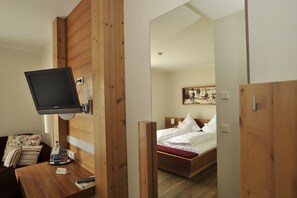 Een hotelkamer met een houten bed, een wandtelevisie, een bureau met een lamp en een spiegel die de kamer weerspiegelt.