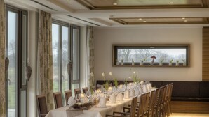 Un espace repas avec une longue table dressée pour le repas, de grandes fenêtres et une vue sur les arbres à l’extérieur.
