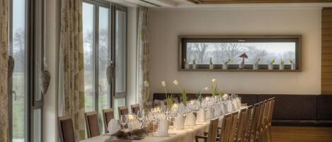 A dining area with a long table set for a meal, large windows, and a view of trees outside.
