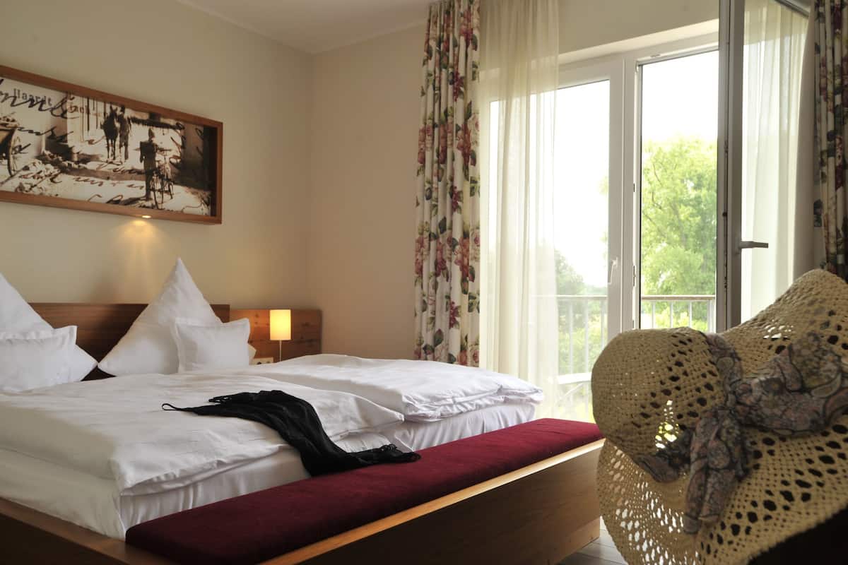 Une chambre d’hôtel avec un grand lit, une tête de lit en bois, une table de chevet avec une lampe, une fenêtre ornée de rideaux à motifs floraux et une vue sur les arbres à l’extérieur.