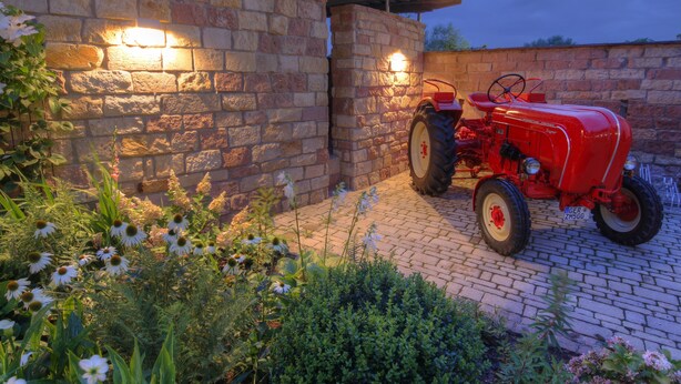 Un tracteur rouge est garé devant un bâtiment en pierre, avec un chemin bordé de briques et des plantes en fleurs.