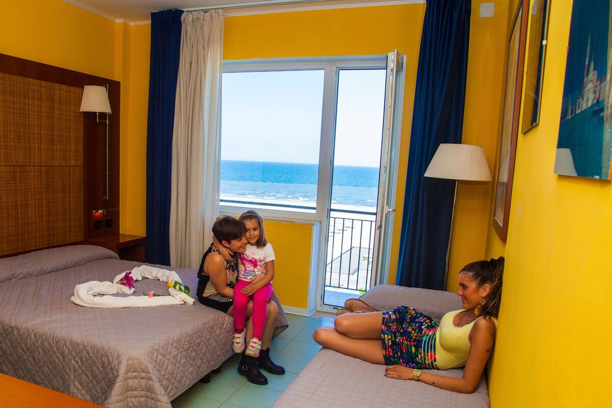 A hotel room with two beds, a balcony with a view of the sea, and a large window.