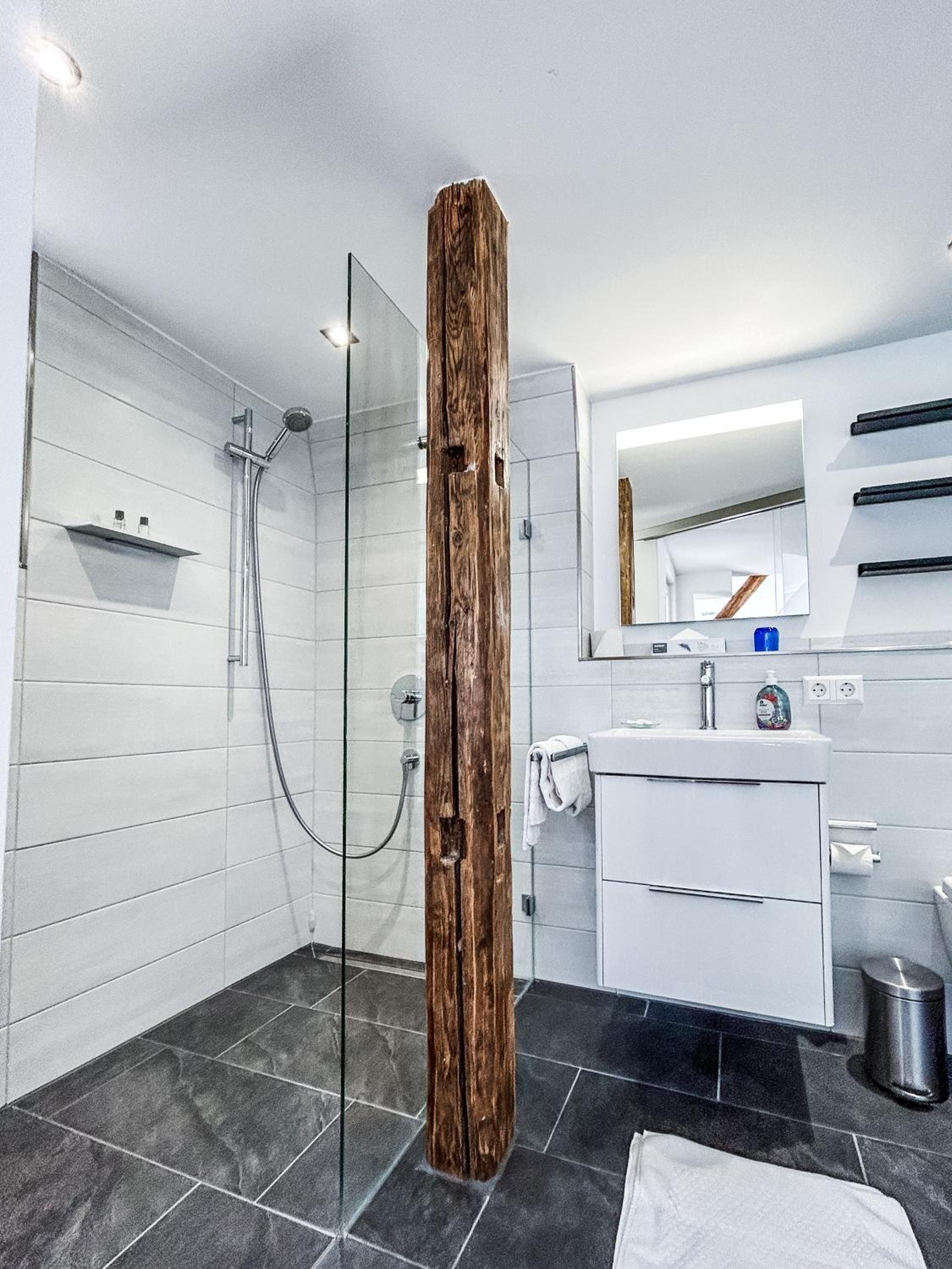 Une salle de bain moderne avec une douche à l’italienne en verre, une colonne décorative en bois, un meuble-lavabo blanc avec un miroir et un sol carrelé de couleur noire.