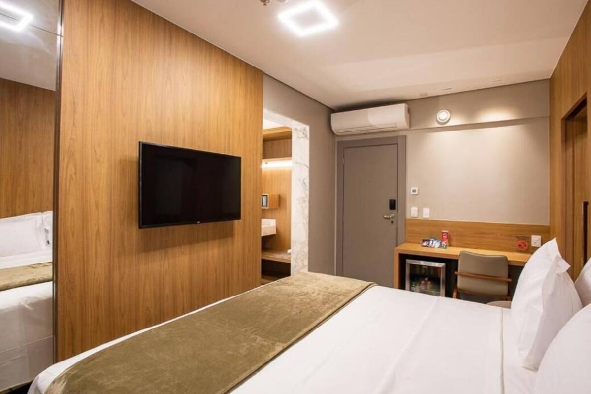 A hotel room with a bed, a TV mounted on the wall, a desk with a chair, and a wardrobe.