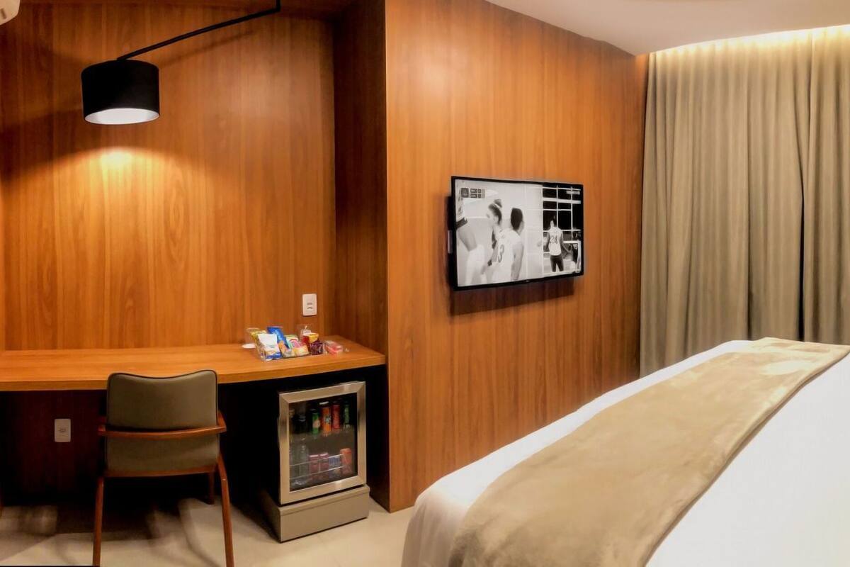 A hotel room with a bed, a desk with a chair, a refrigerator, a television mounted on the wall, and a curtain.