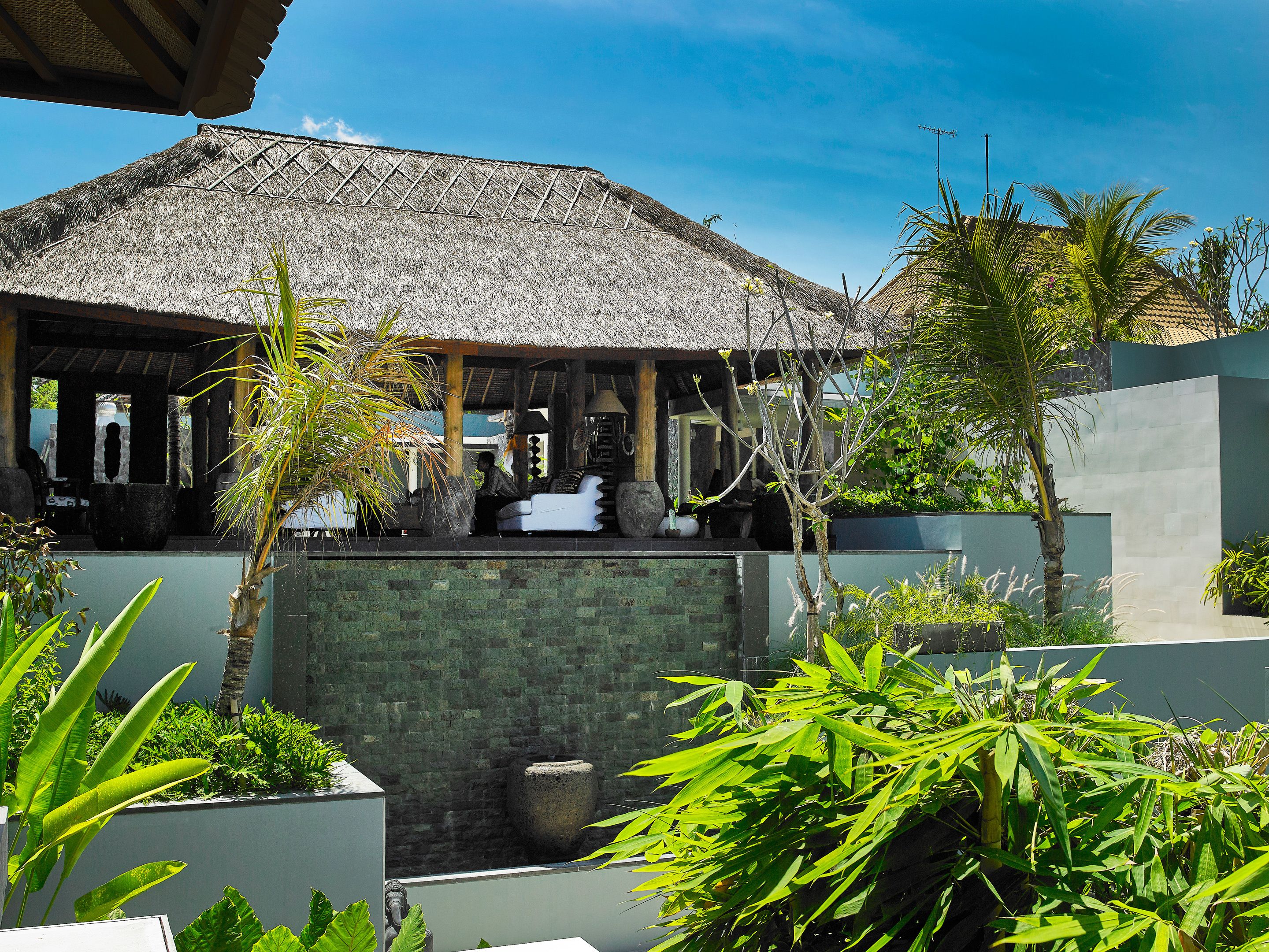 A thatched-roof structure with a covered seating area, surrounded by greenery and a stone wall.