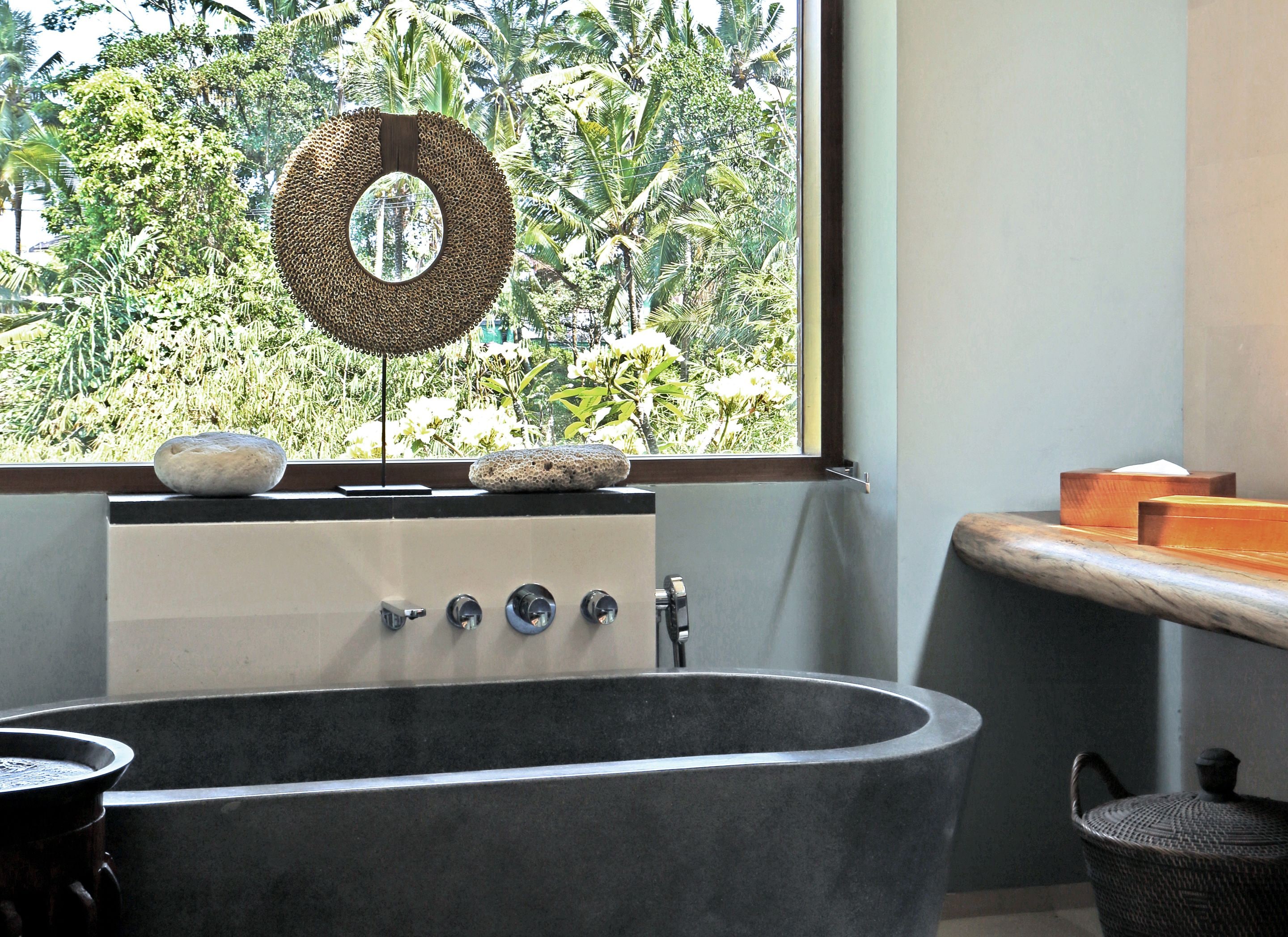 Un baño con una bañera independiente, una ventana que ofrece vistas a follaje tropical y un perchero con una cesta tejida.