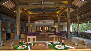 A dining area with a long wooden table set for a meal, surrounded by wooden chairs and a sofa. - The Purist Villas & Spa (Ubud)