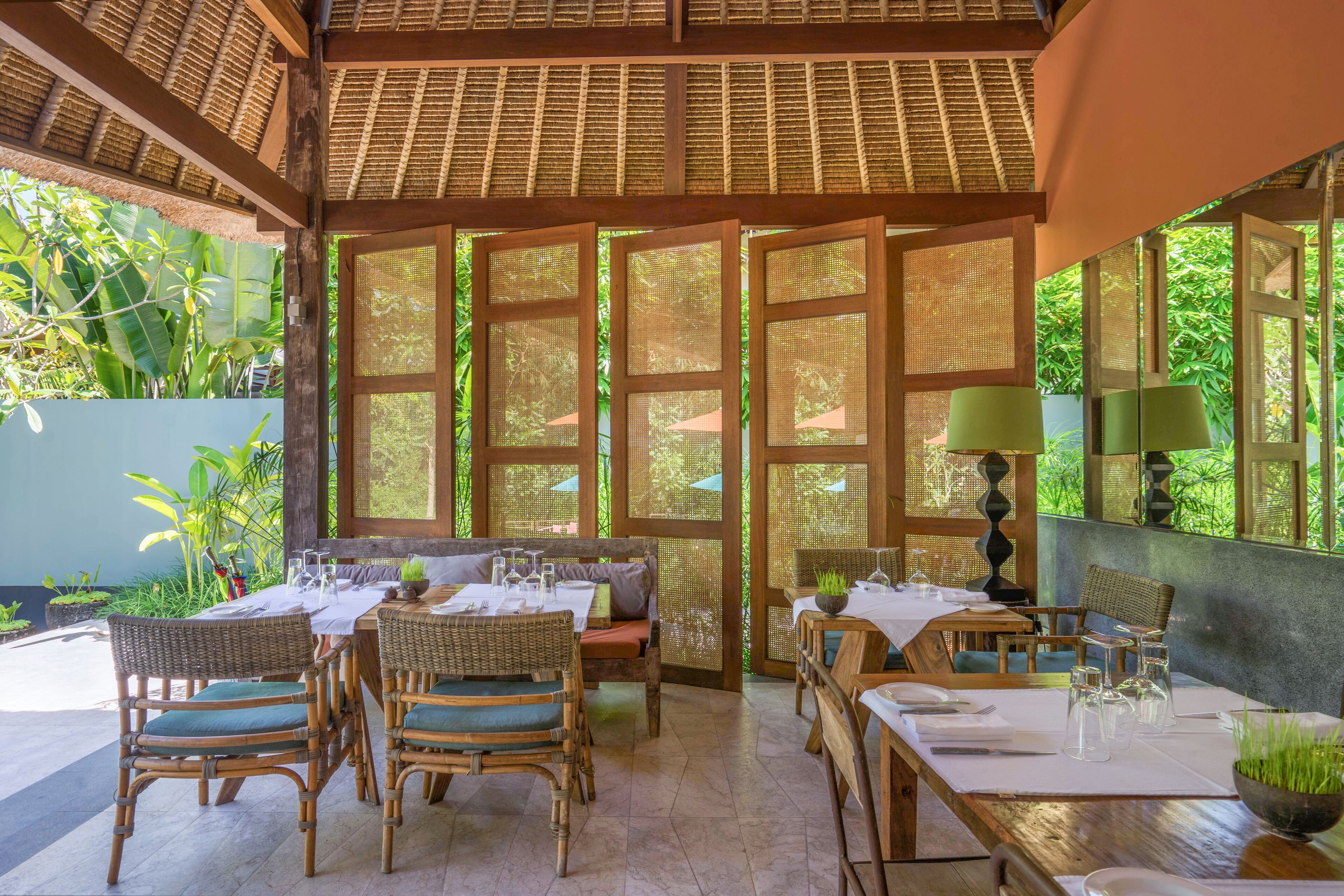 An indoor dining area with wicker furniture, a high ceiling with a woven pattern, and large windows allowing natural light.
