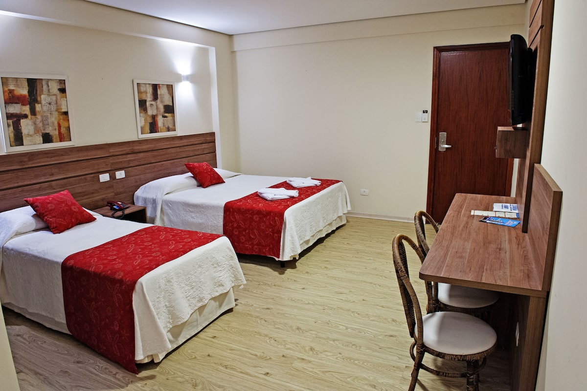 A hotel room with two beds, each with a red and white bedspread, a wooden headboard, a small desk with a chair, and a television mounted on the wall.