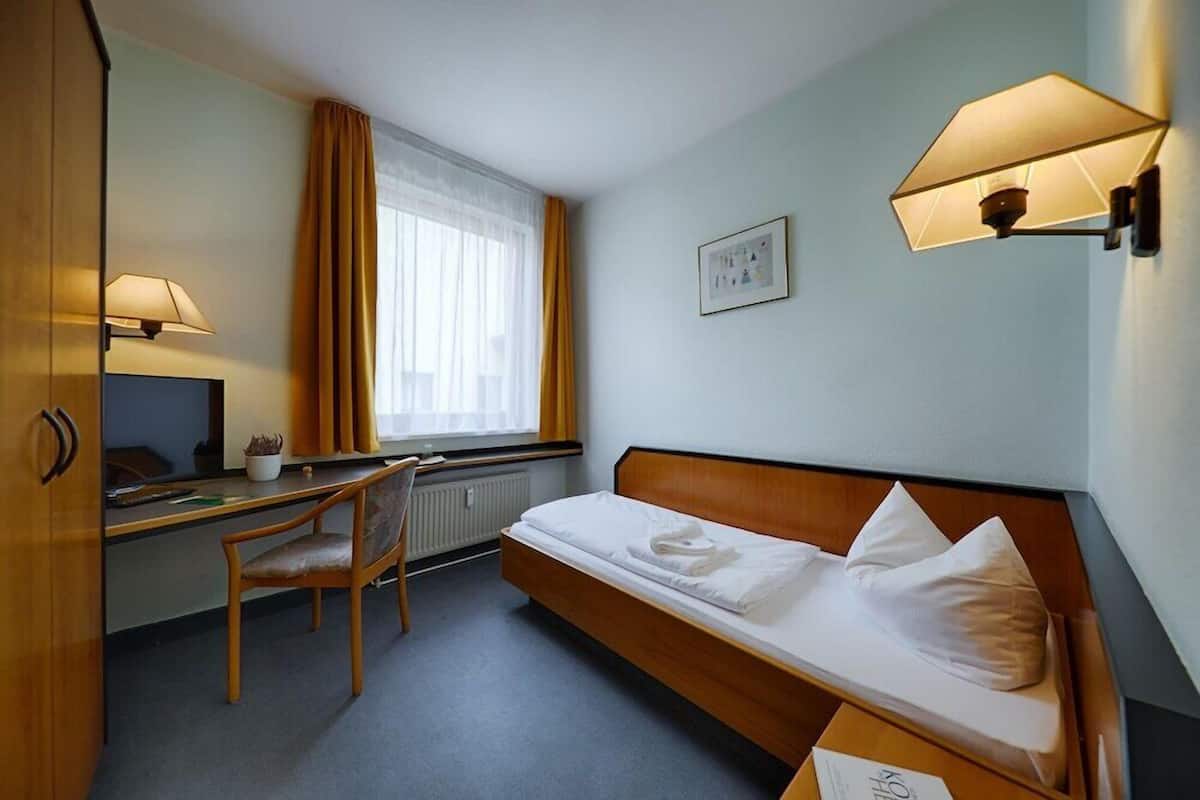 A hotel room with a bed, a desk, a chair, a window with curtains, a wall-mounted lamp, and a framed picture.