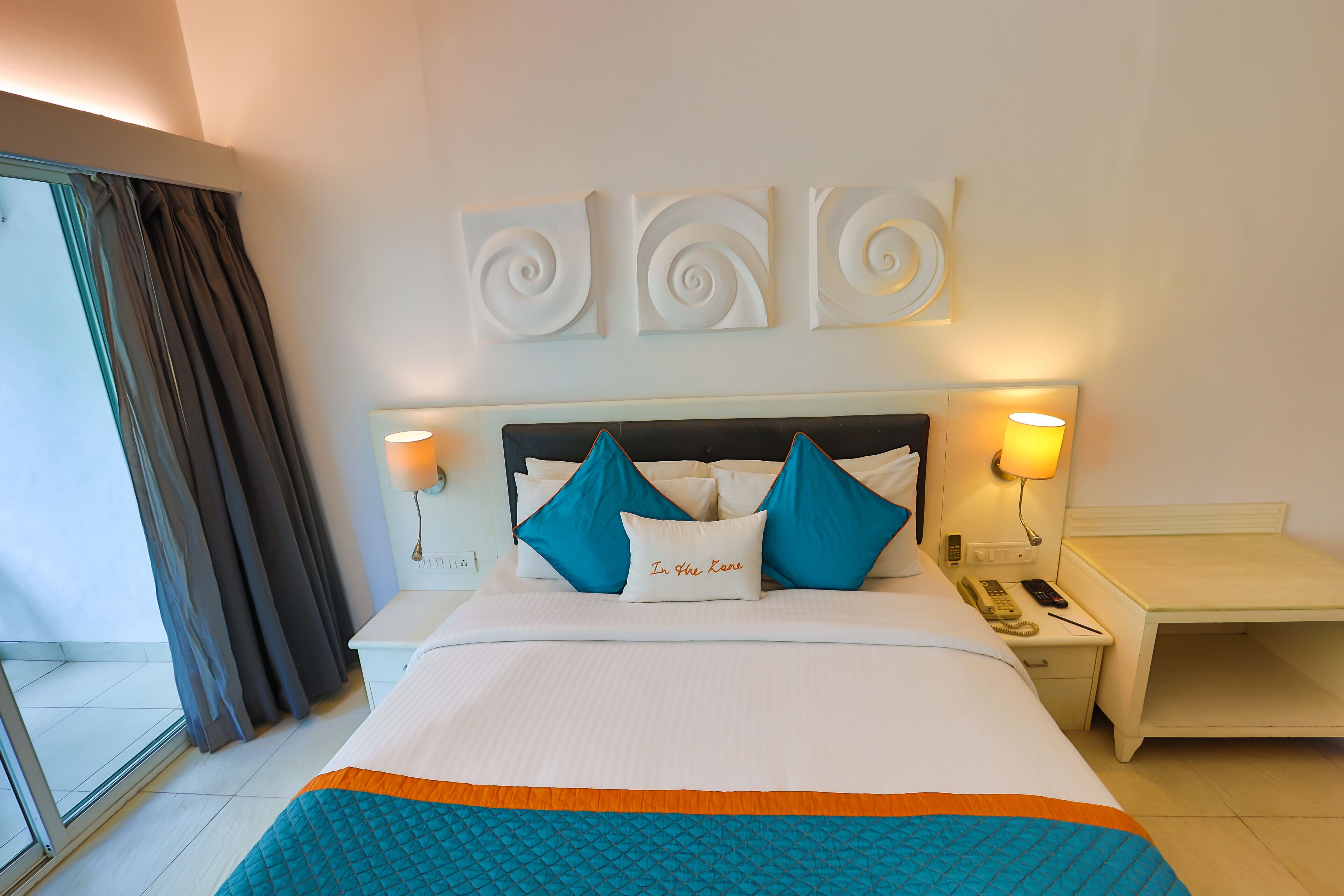 Una habitación de hotel con una cama, dos mesitas de noche, una lámpara y una obra de arte colgada en la pared.