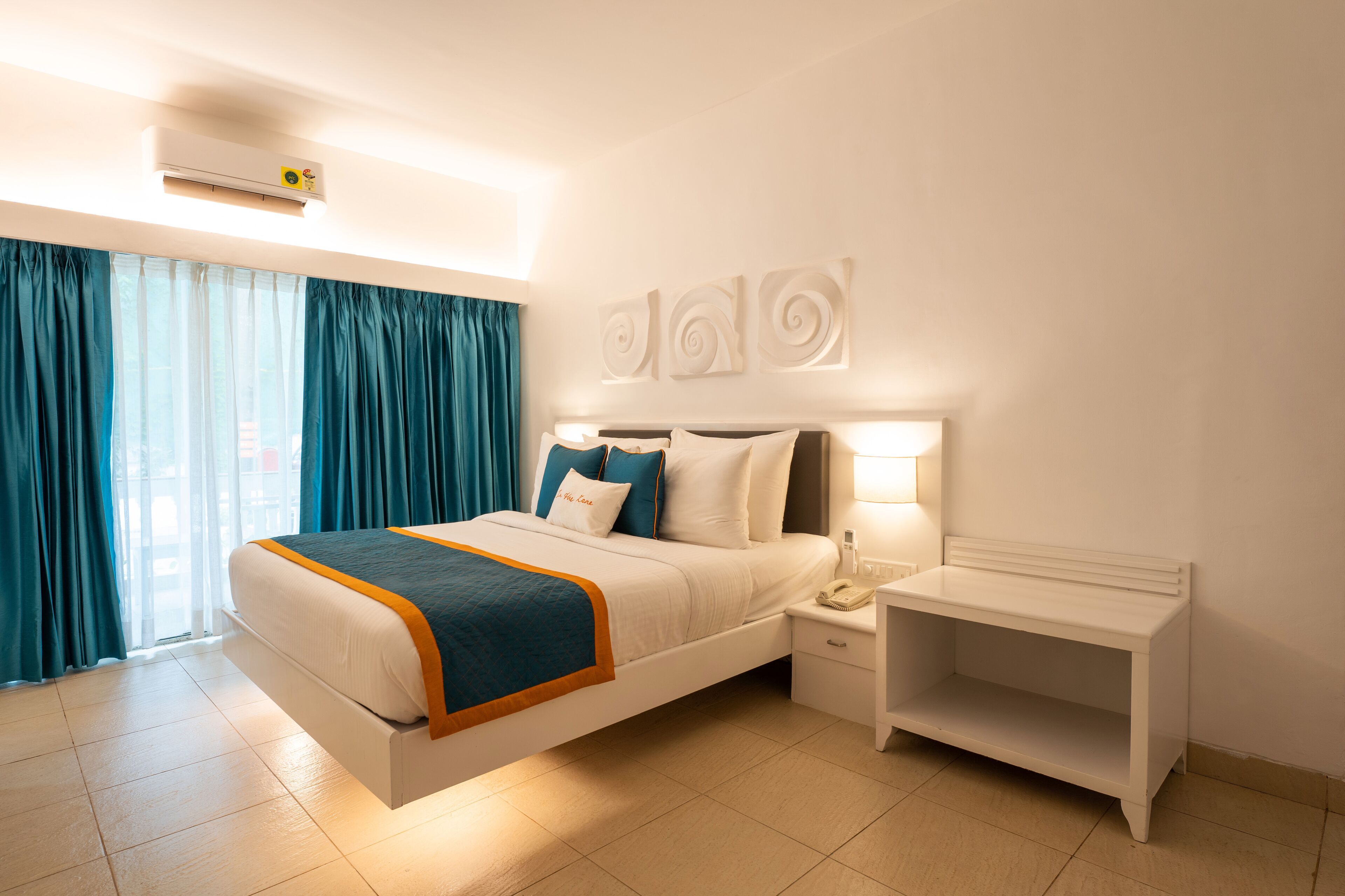 Una habitación de hotel moderna con una cama grande, una mesita de noche y un ventanal con cortinas.