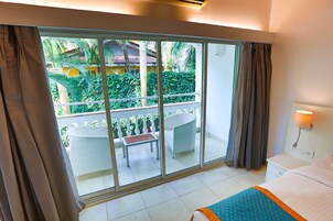 Habitación de hotel con balcón, cama, mesita de noche y vistas a un jardín y un edificio.