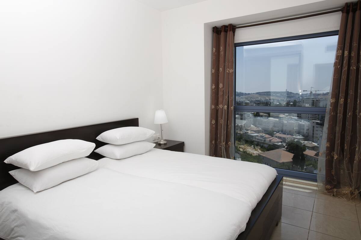 Habitación de hotel con una cama grande, una mesita de noche con lámpara y vistas a un paisaje urbano a través de un ventanal.