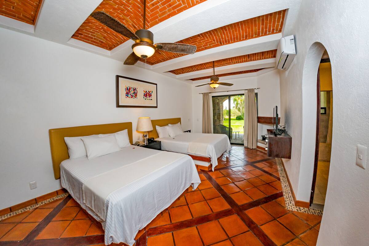 Habitación de hotel con dos camas, ventilador de techo, una obra de arte enmarcada y vistas al exterior.