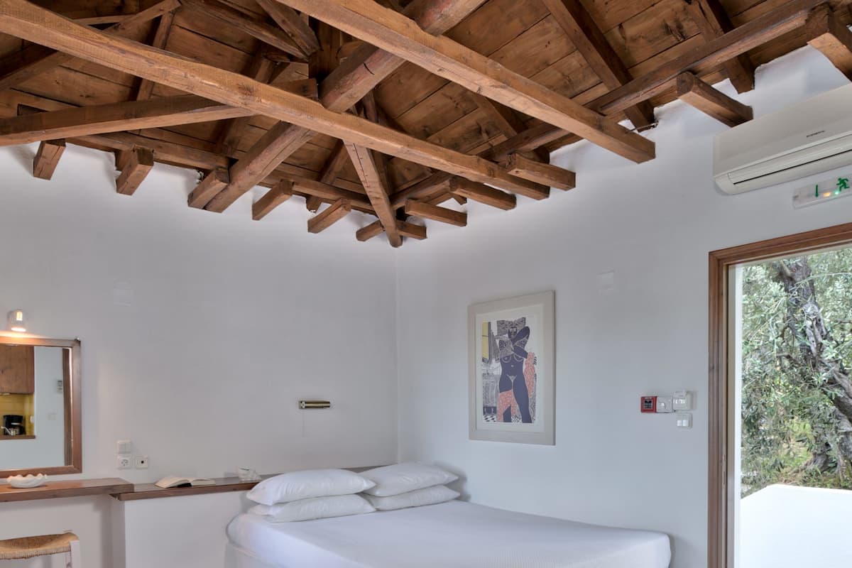 A bedroom with a wooden ceiling, a bed, a desk with a chair, and a window with a view of trees.