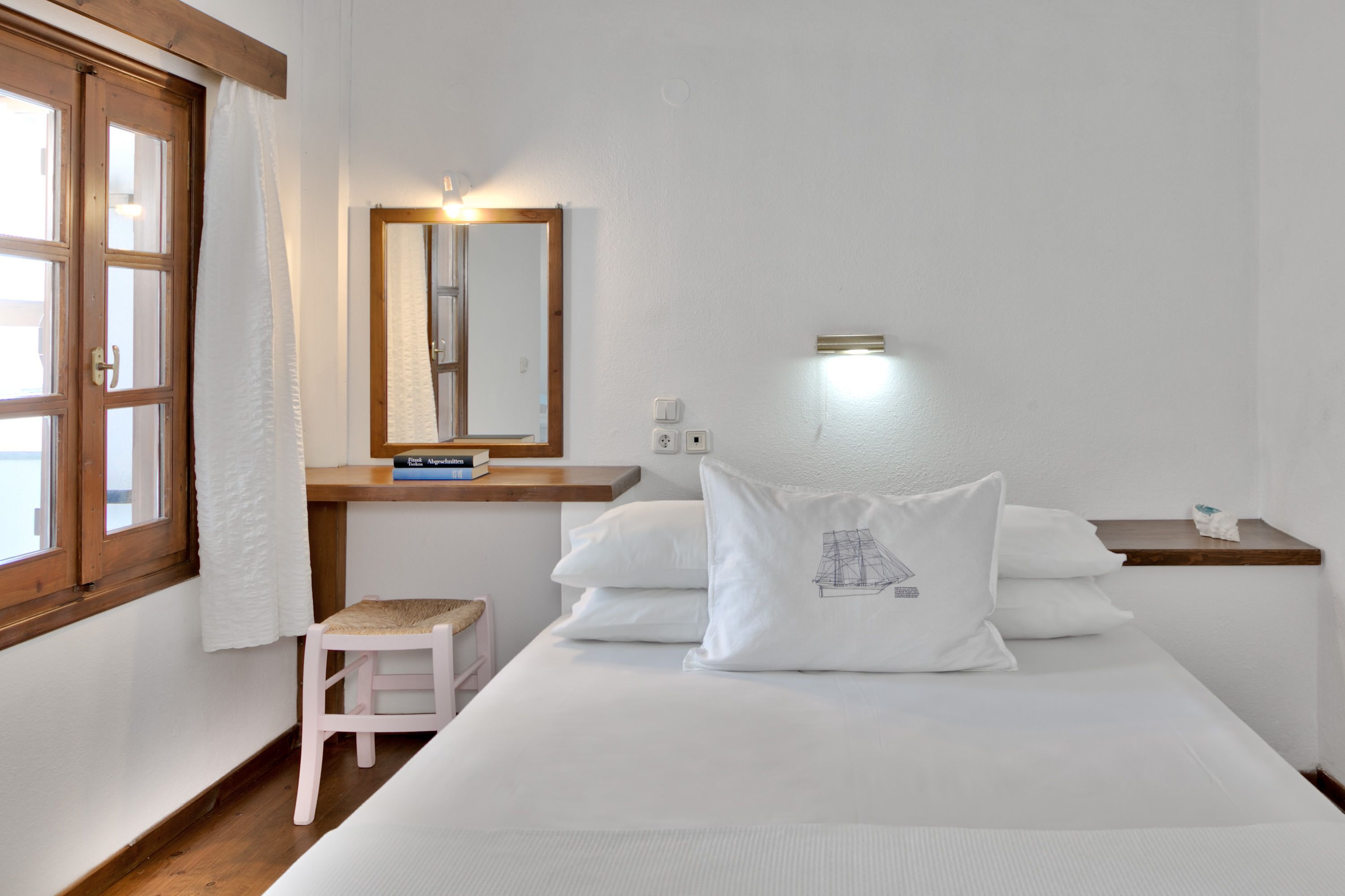 Un dormitorio con cama, un taburete pequeño, una mesita de noche, un espejo y una ventana con cortina.