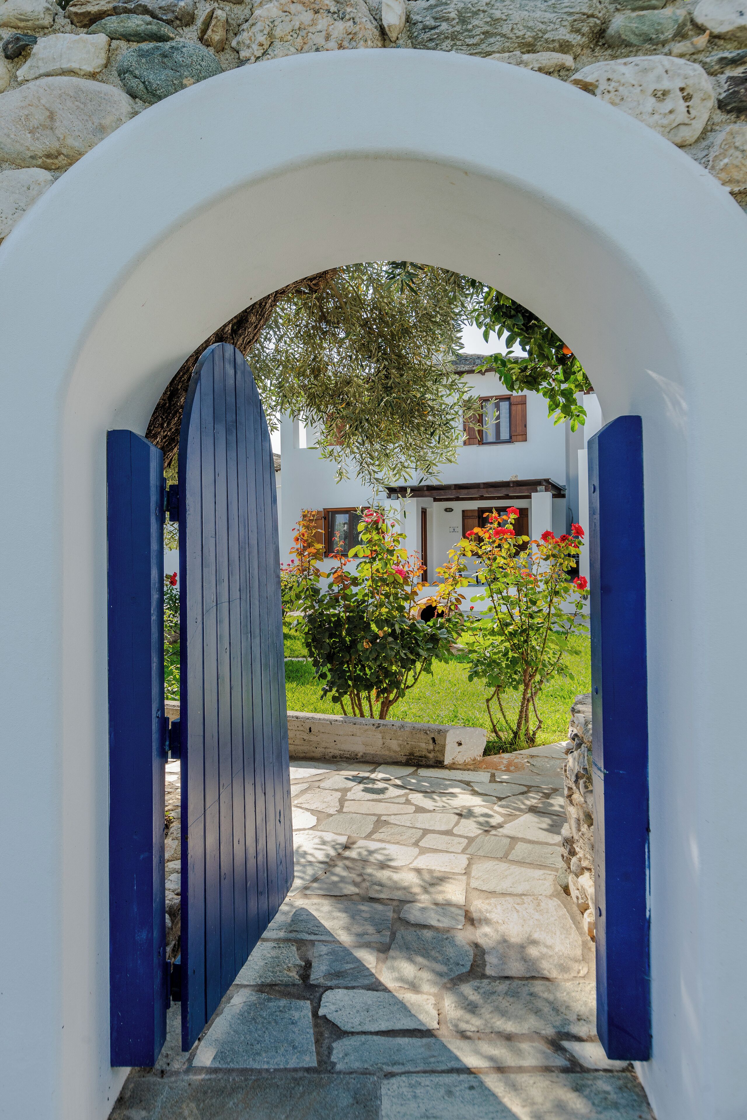 An arched doorway leading to a stone pathway, with a building and blooming flowers in the background.