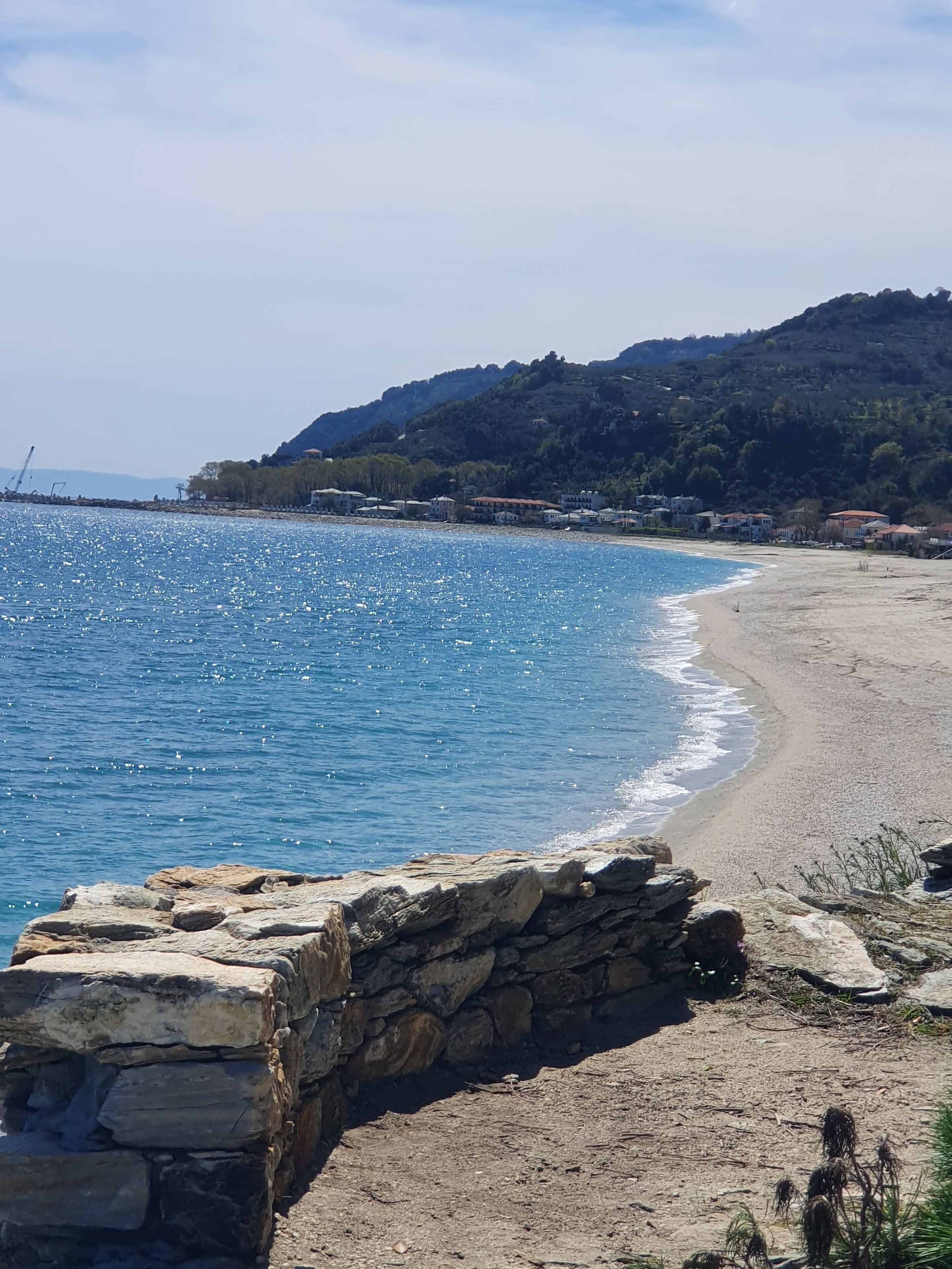 A coastal beach with a stone wall in the foreground, a clear blue sea, and a hillside with buildings in the background.