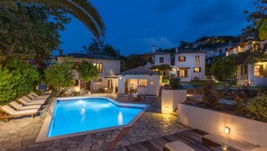 A swimming pool with lounge chairs, a stone patio, and a hillside view of houses at dusk. - Aeolos Hotel & Villas -Pelion (Zagora-Mouresi)