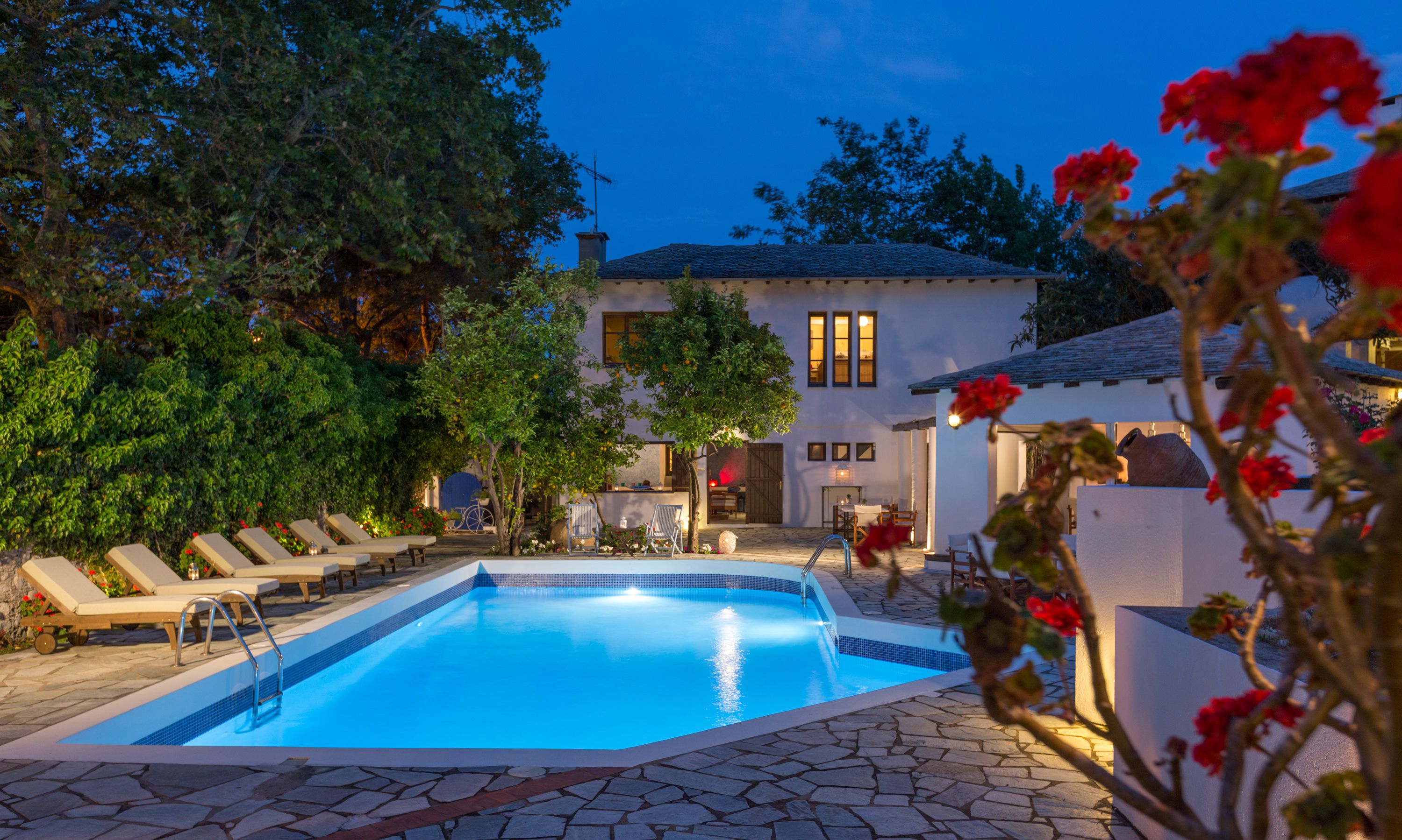 Une piscine entourée de chaises longues, une terrasse dallée de pierres et un bâtiment blanc doté de nombreuses fenêtres.