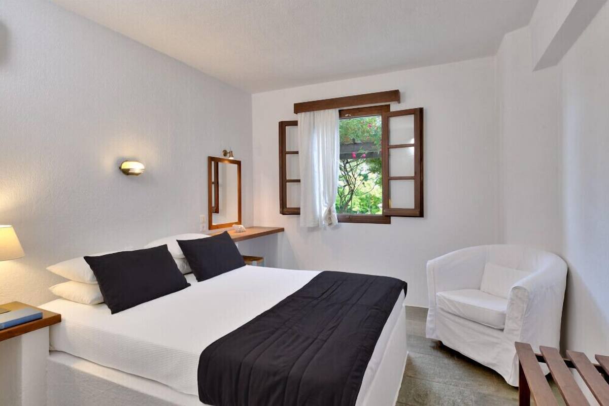 A hotel room with a bed, a chair, a nightstand, a lamp, and a window with a view of greenery.