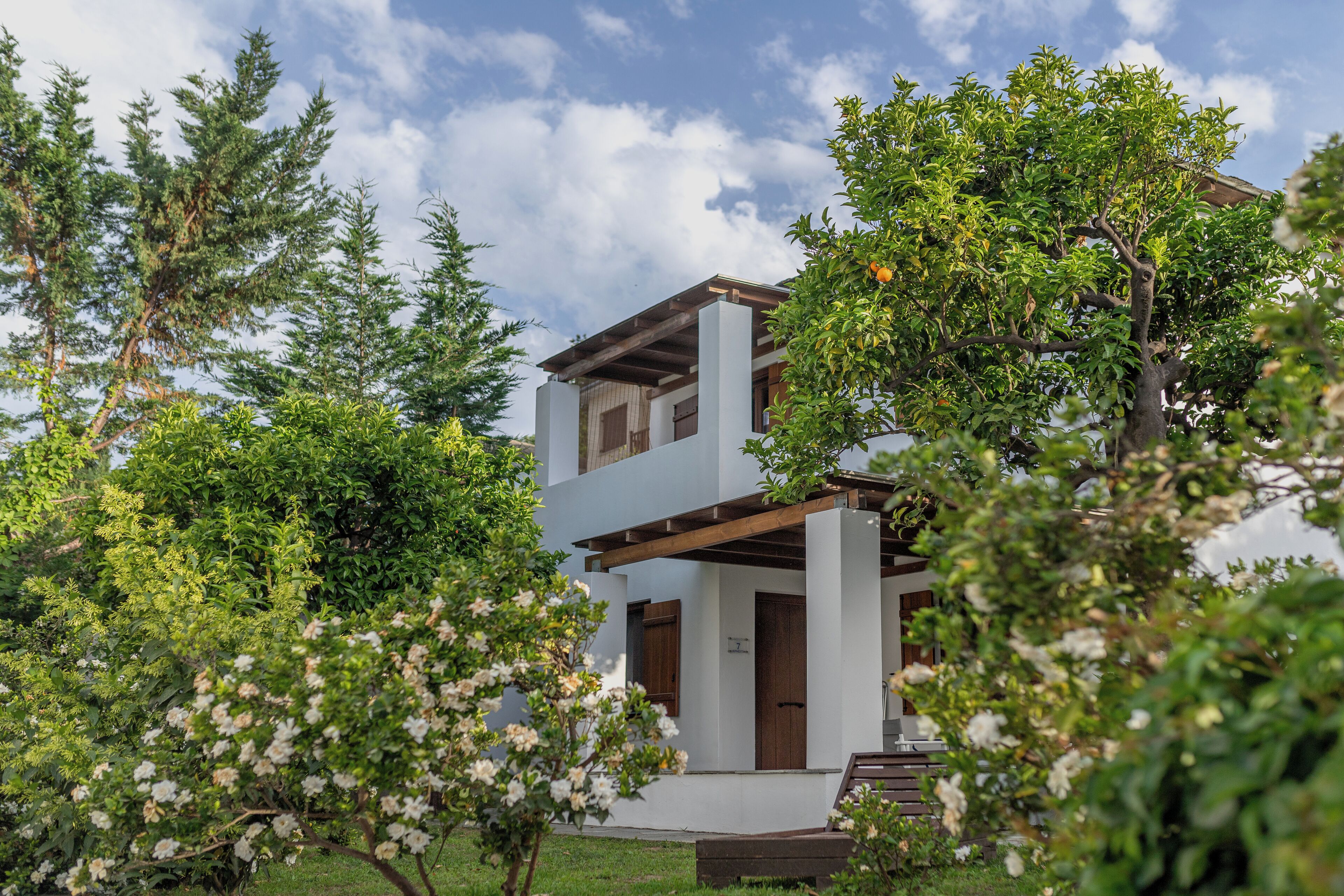 A two-story building with a white facade and wooden beams, surrounded by lush greenery and flowering trees.