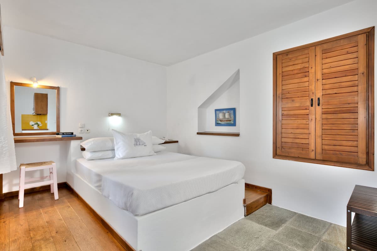 A bedroom with a bed, a wooden wardrobe, a small stool, a nightstand, and a wall-mounted shelf with a framed picture.