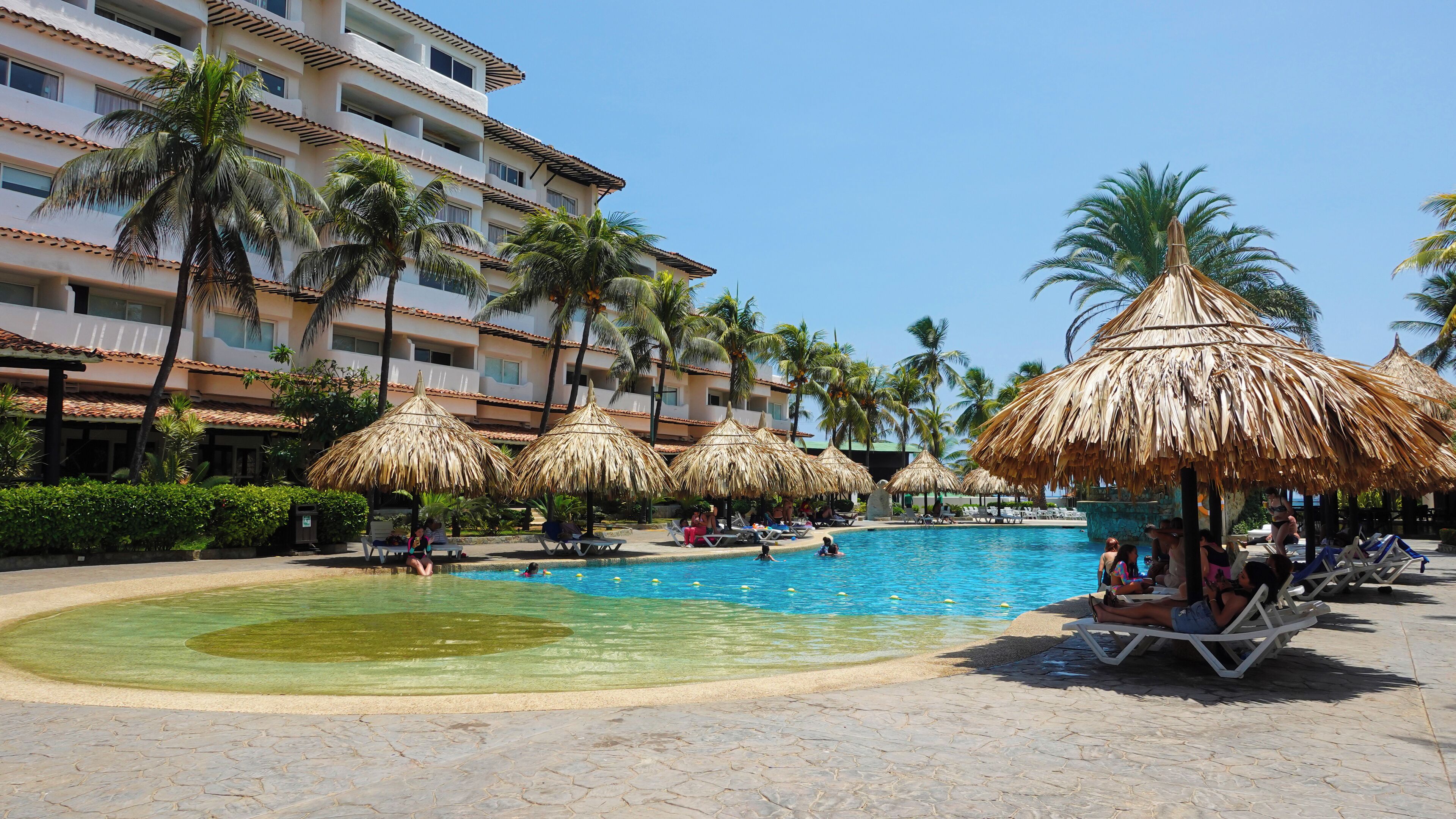 A resort pool area with clear water, palm trees, and thatched umbrellas.