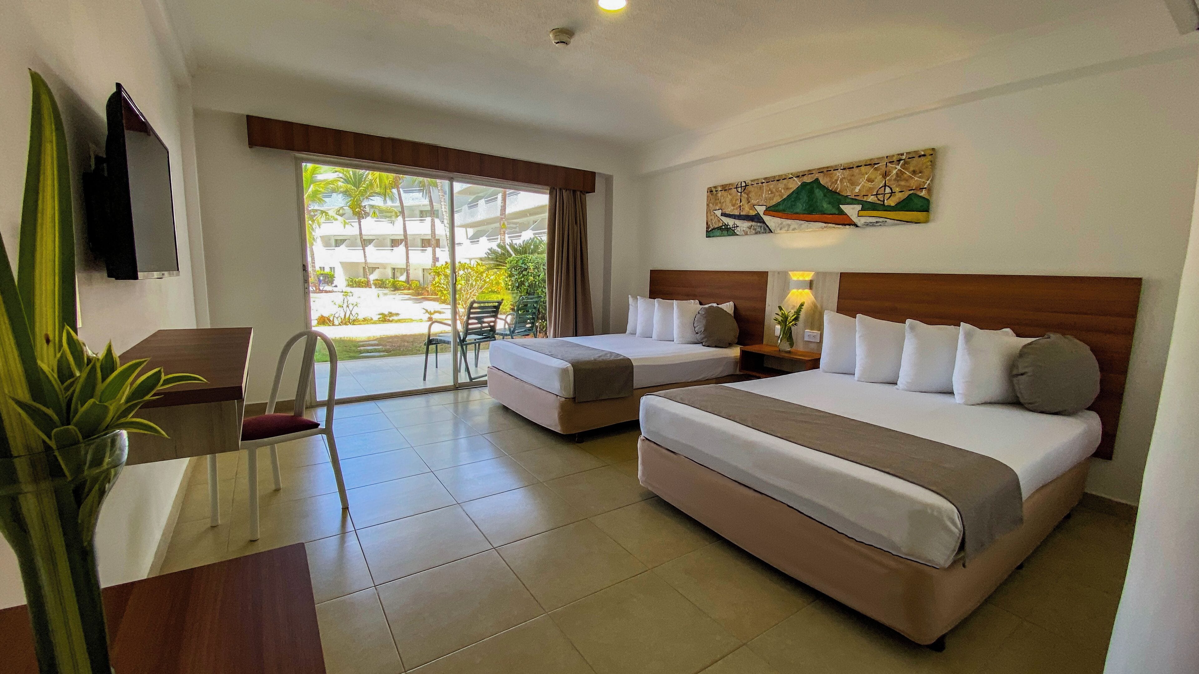 Habitación de hotel con dos camas, un escritorio, una silla, televisión y ventana con vista a palmeras y edificios.