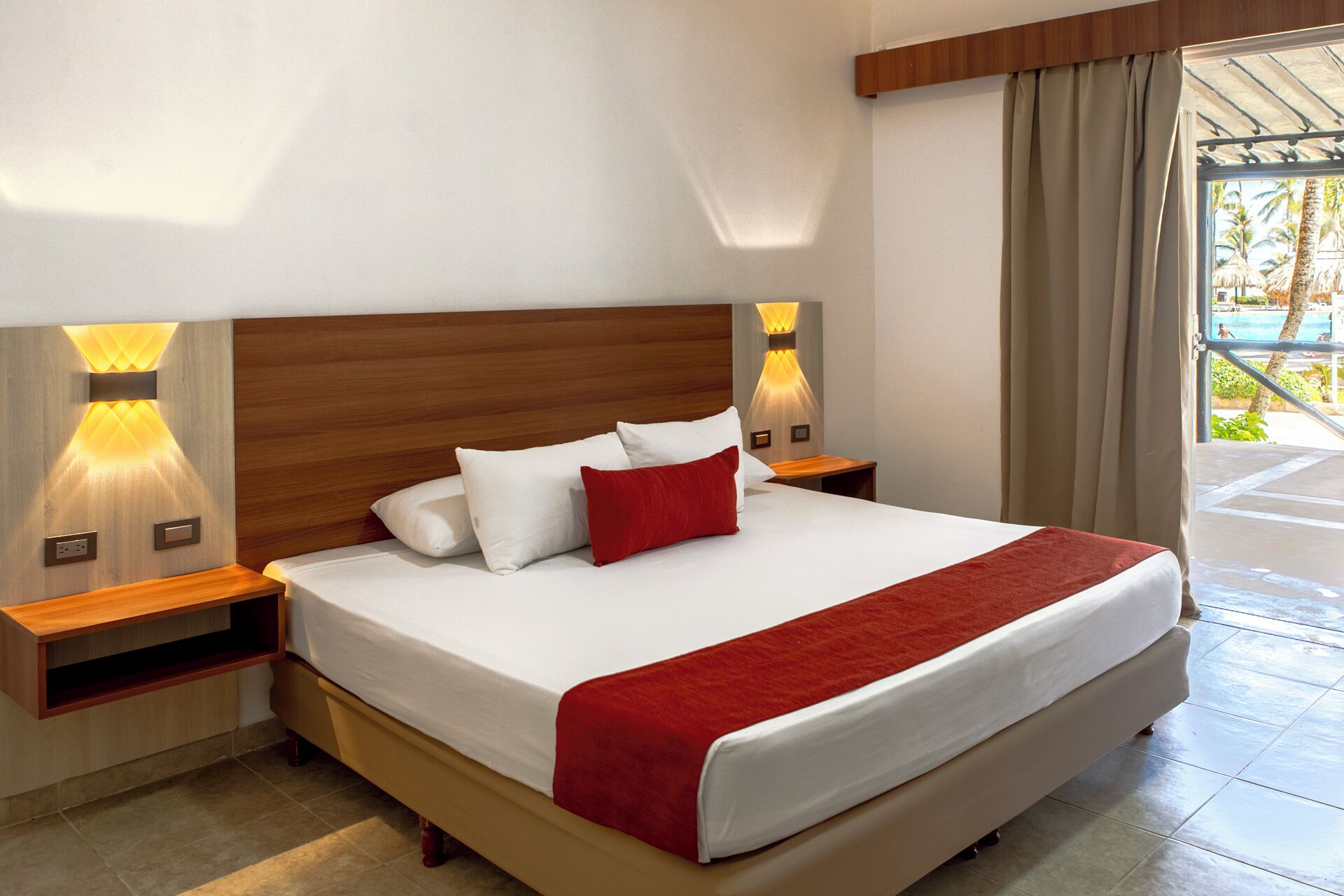 A hotel room with a large bed, wooden headboard, two bedside tables with lamps, and a view of a pool area.
