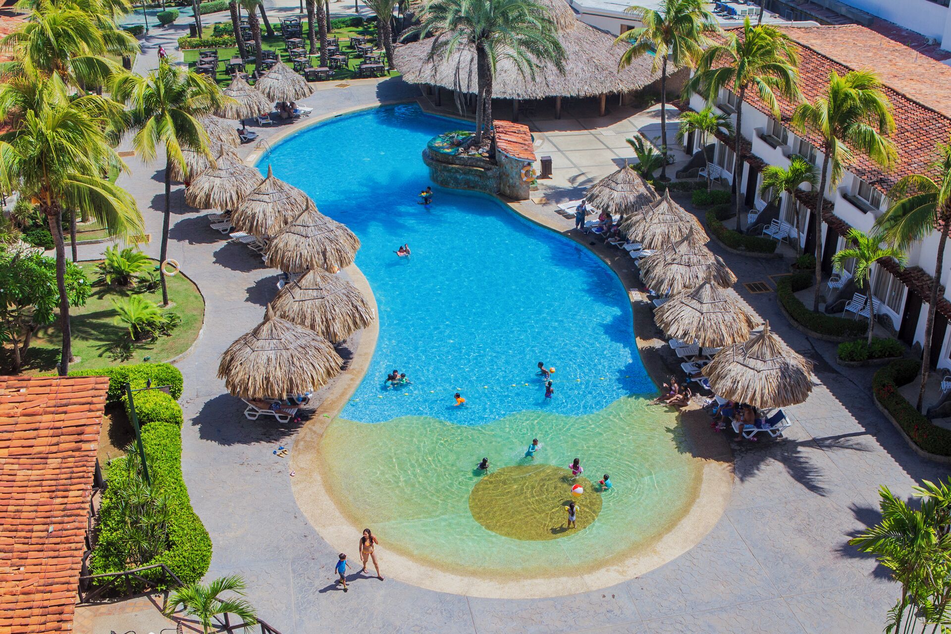 A resort with a large swimming pool surrounded by palm trees and thatched umbrellas.