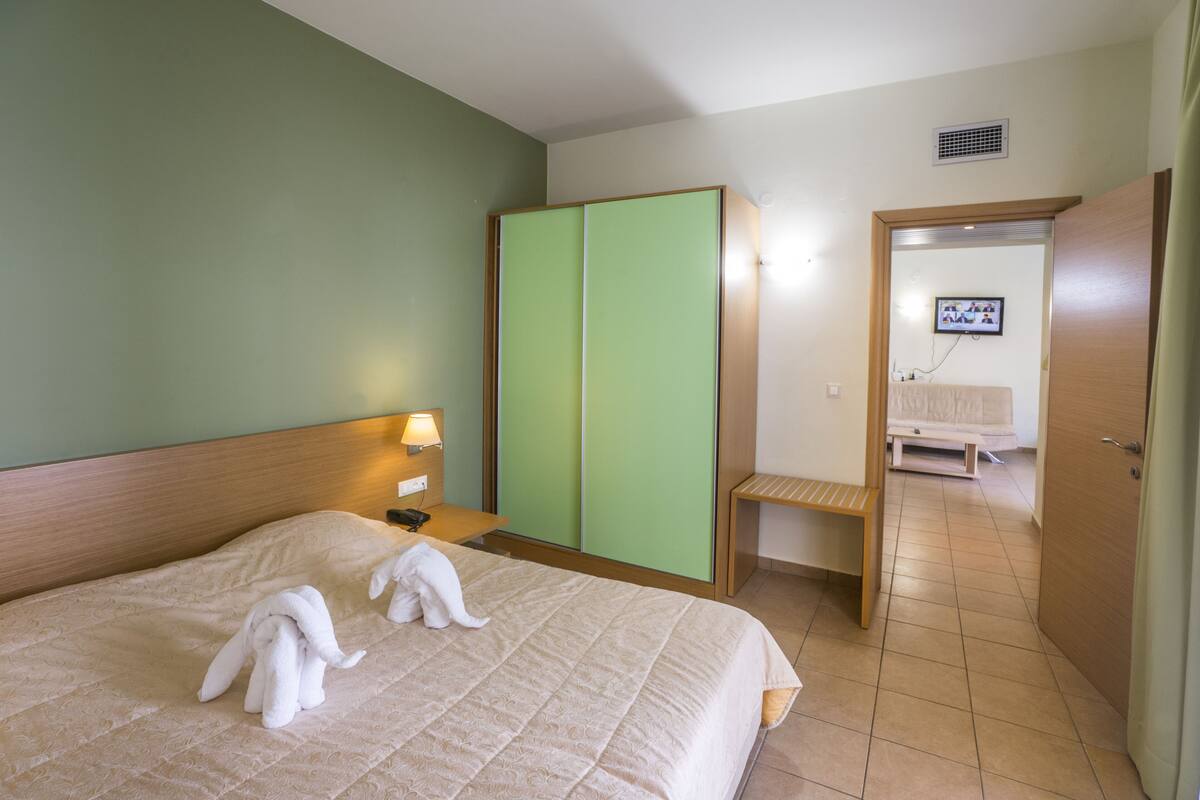 Une chambre d’hôtel comprenant un lit, une table de chevet, une armoire verte, un banc en bois et une porte donnant sur une autre pièce.