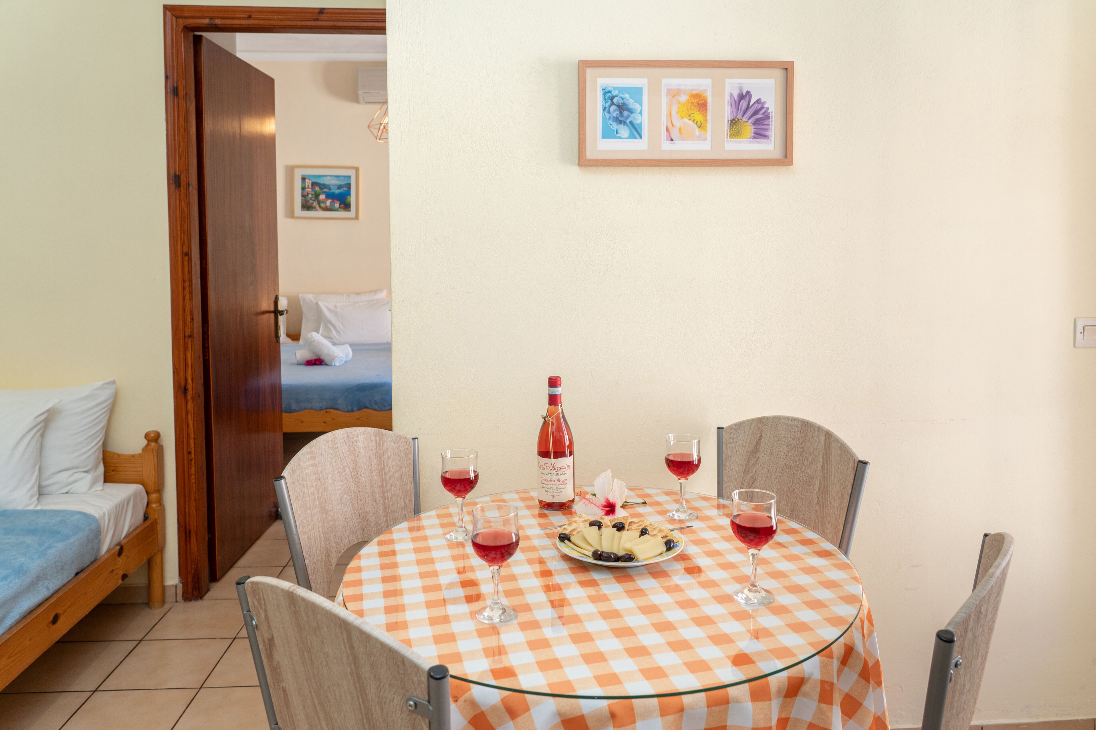 Une pièce avec un lit, une table à manger recouverte d’une nappe à carreaux, deux chaises, une bouteille de vin et une assiette de nourriture.