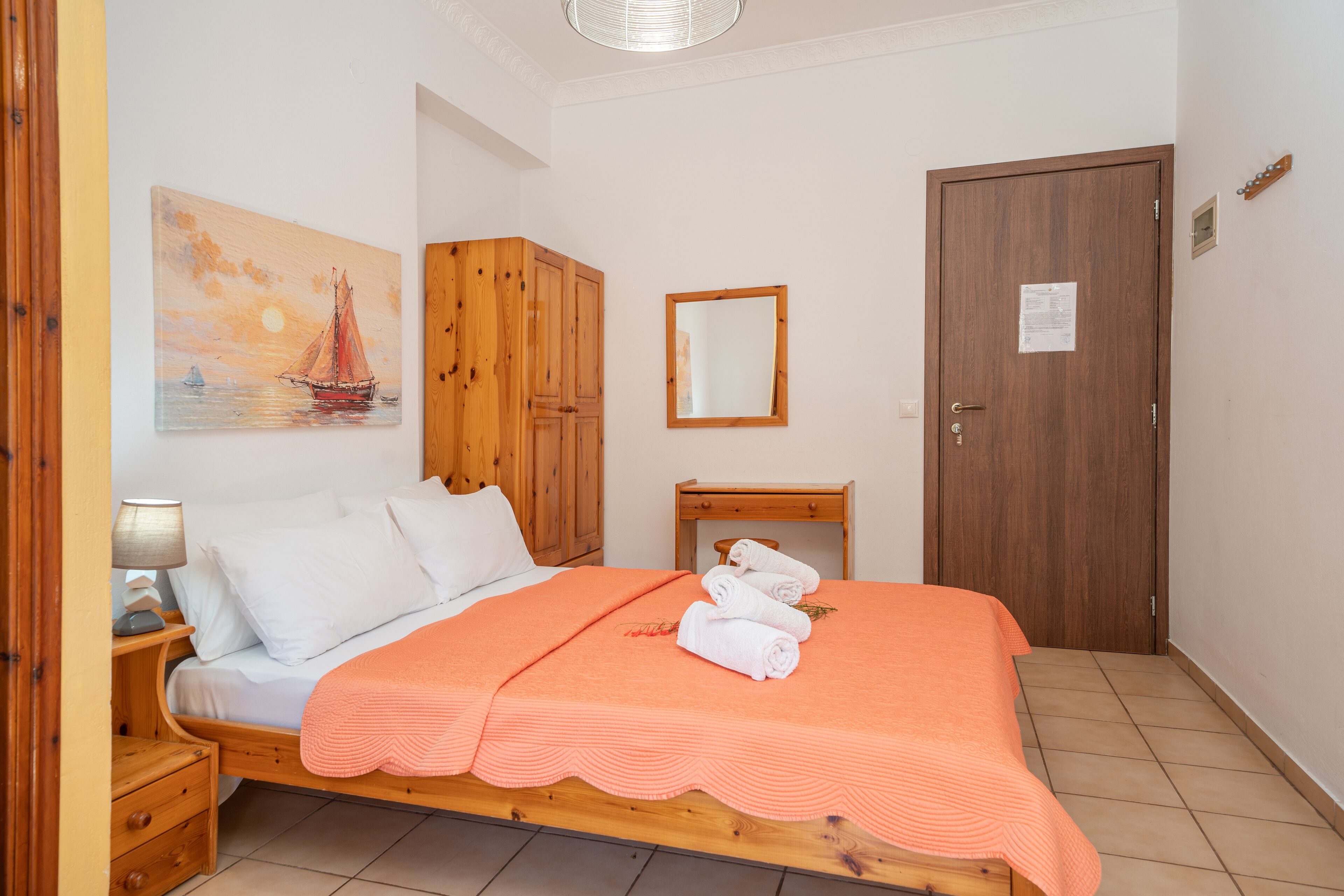 Un dormitorio con cama de madera, ropa de cama blanca, un cuadro de un velero, un armario de madera, un espejo y una puerta con un papel adherido.