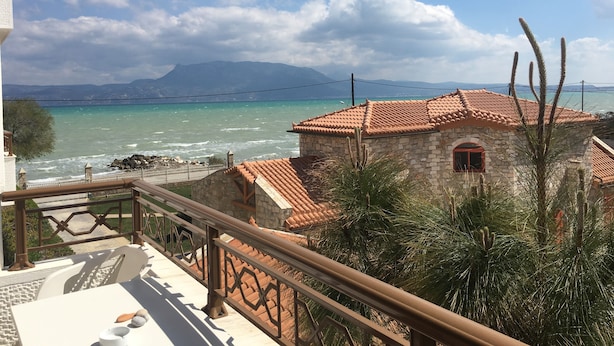 A stone building with a tiled roof, a balcony with a railing, and a view of the sea and mountains in the background.