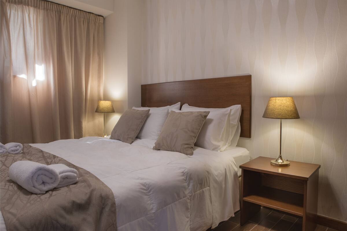 Ein Hotelzimmer mit einem aufgeräumten Bett, zwei Nachttischlampen, einem Holznachttisch und einem Fenster mit Vorhängen.