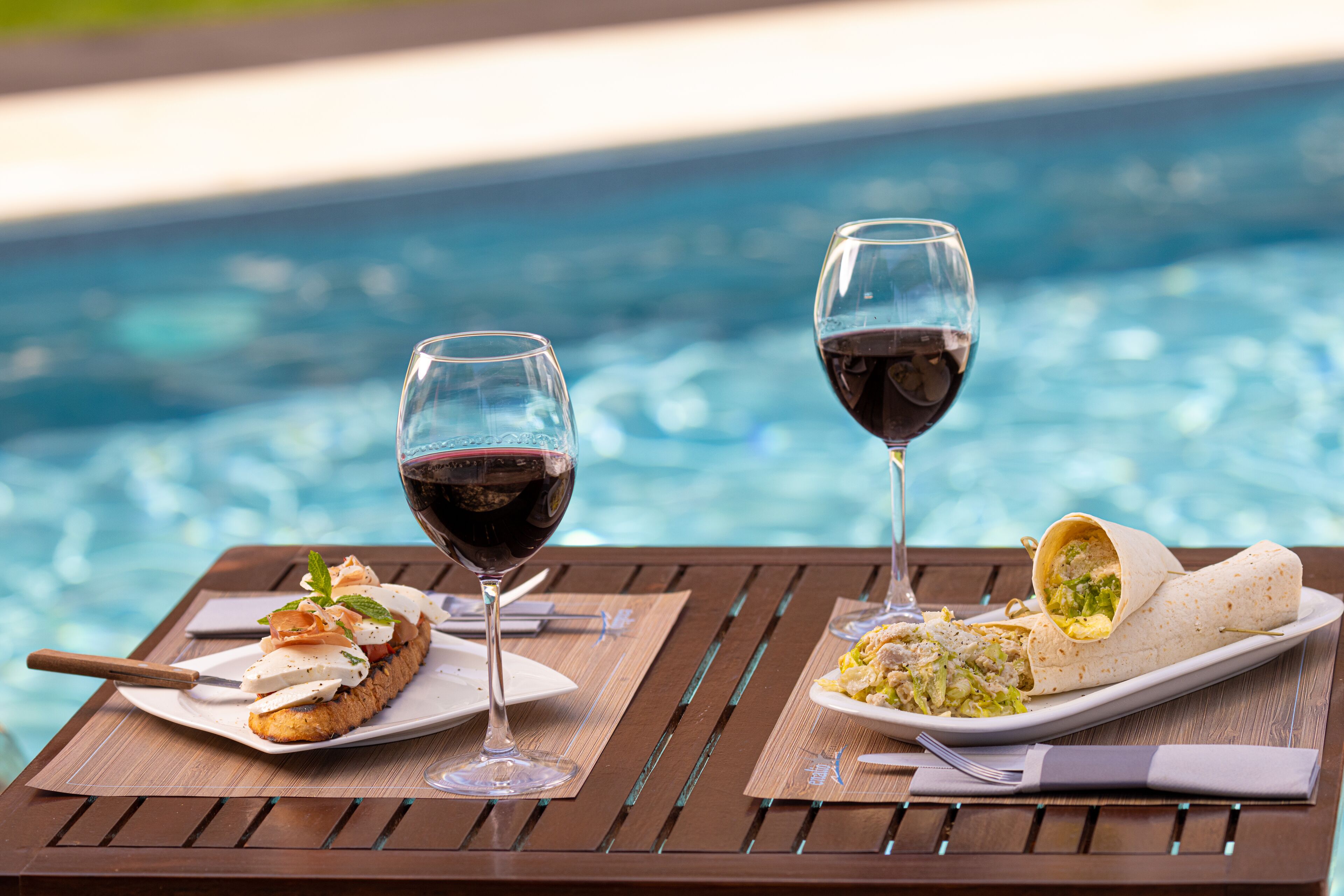 A dining table by a pool with a plate of food and two glasses of red wine.