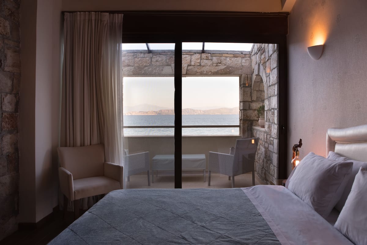 Ein Hotelzimmer mit einem Bett, einer Sitzecke mit Stühlen, einem Tisch und Meerblick durch ein großes Fenster.