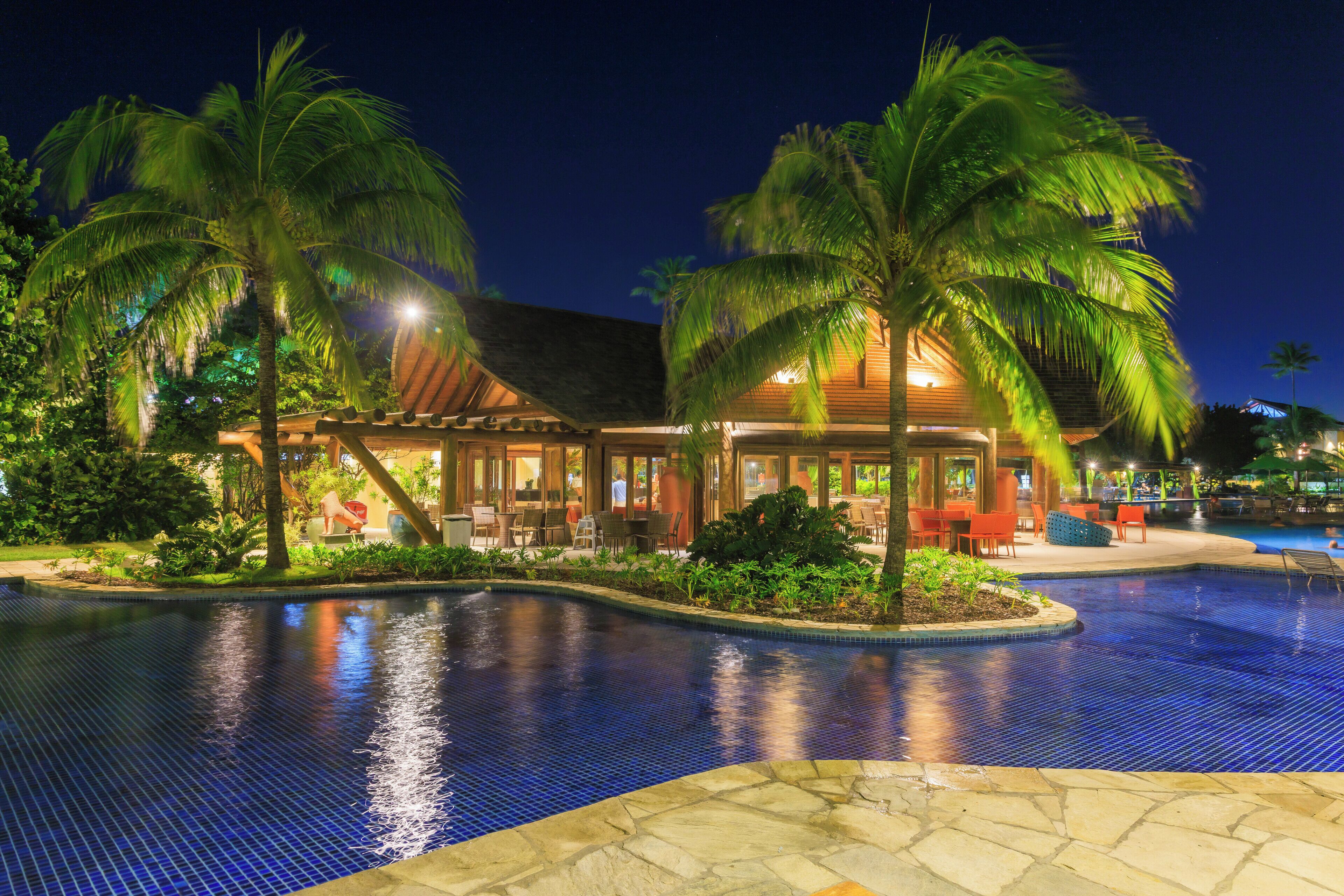 A pool area with a tiled edge, palm trees, and a building with outdoor seating.