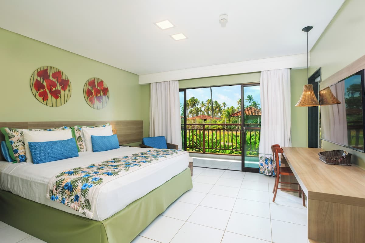 A bedroom with a large bed, a desk, and a balcony with a view of palm trees.