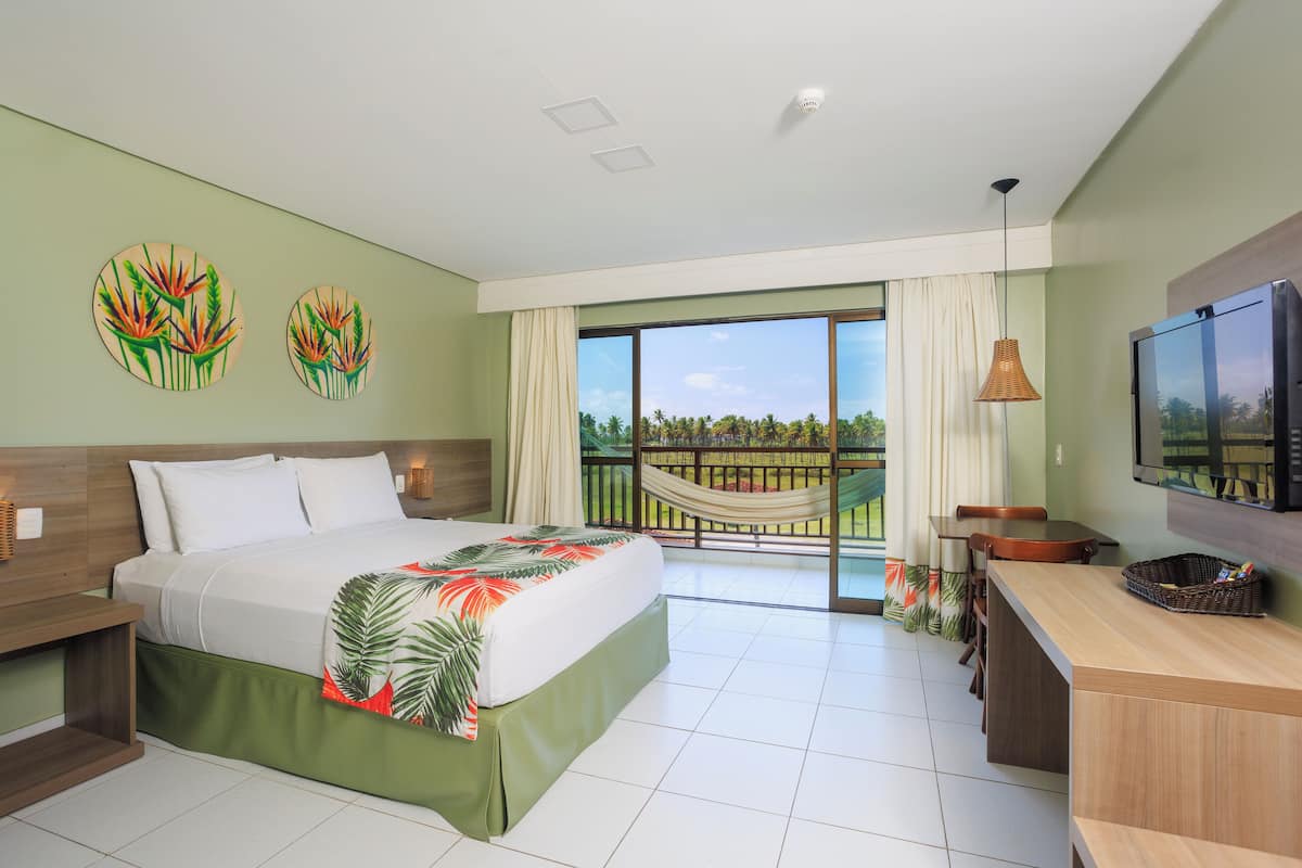 A hotel room with a bed, a desk, a TV, a balcony with a hammock, and a view of palm trees.