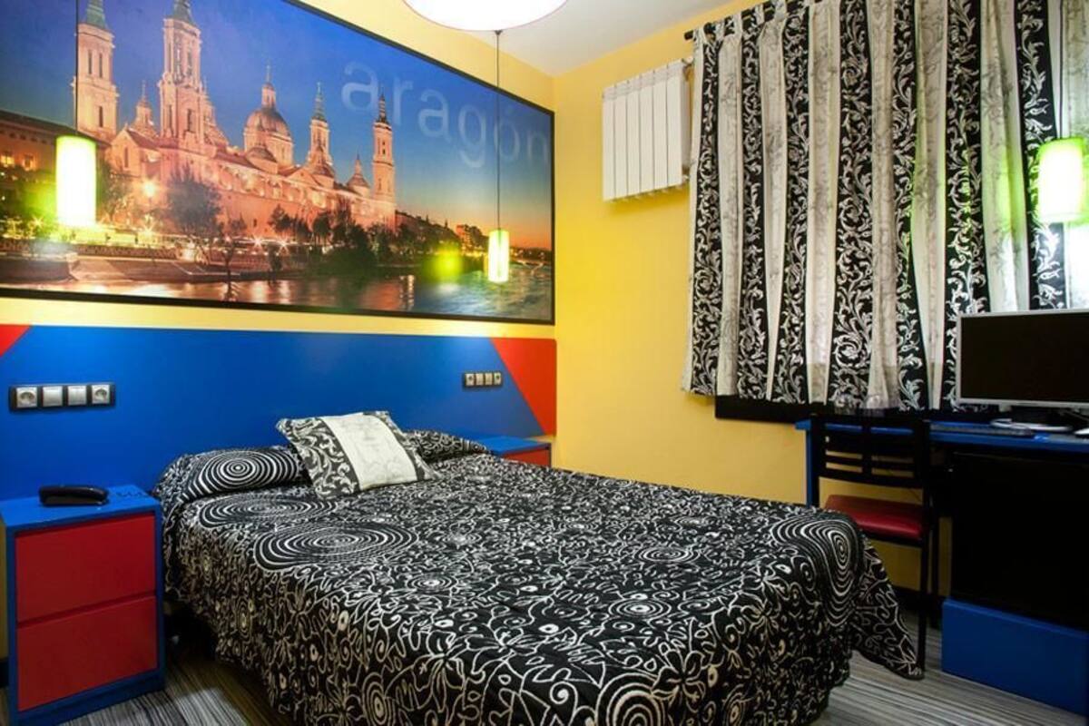 A hotel room with a bed, a desk, a chair, a TV, a wall with a cityscape painting, and a window with curtains.