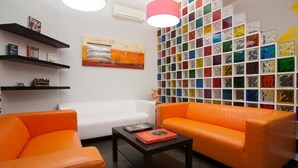 A modern living room with orange sofas, a white couch, and a coffee table. There are shelves with books and a colorful mosaic wall. - JC Rooms Santa Ana (Madrid)