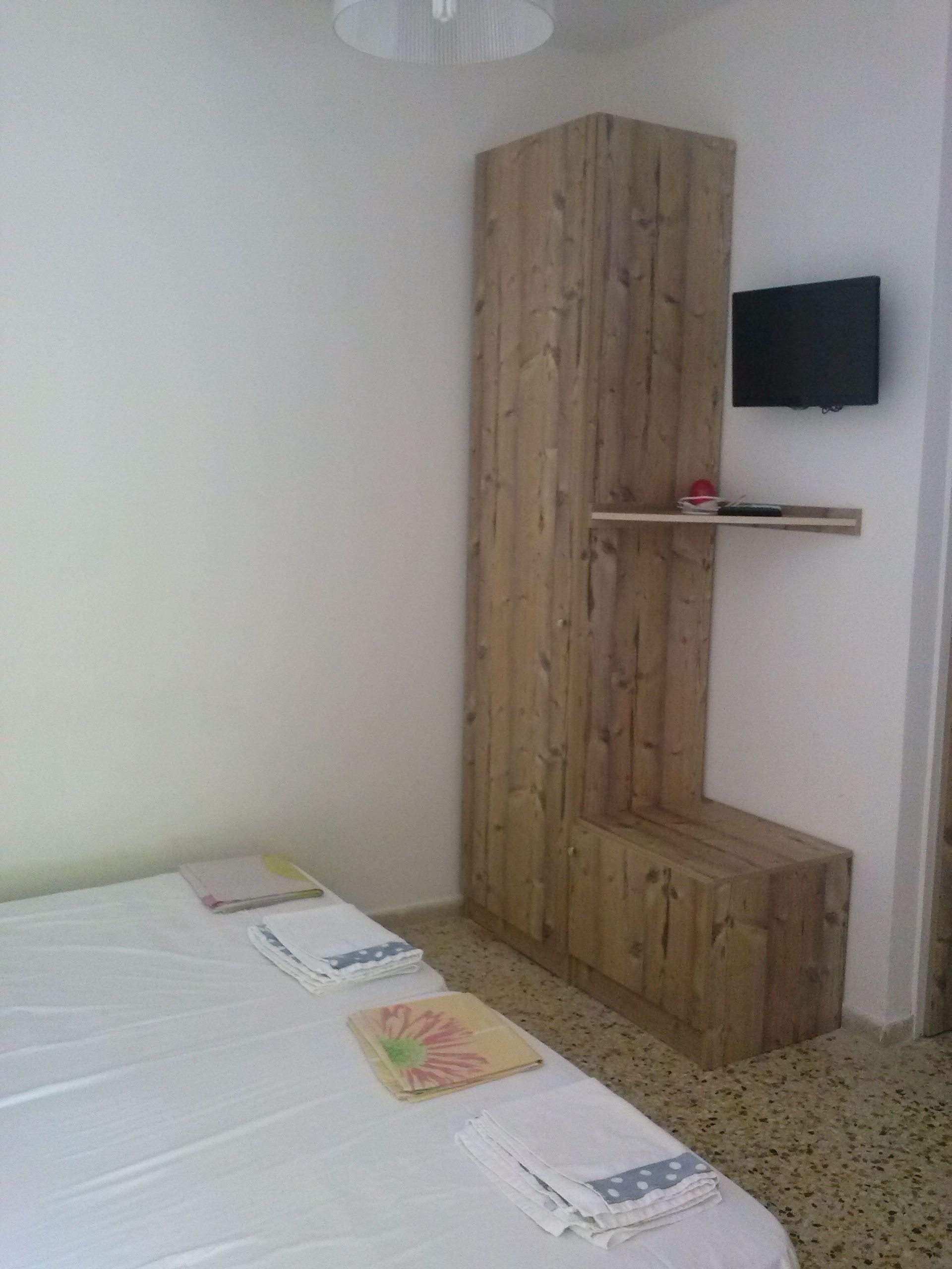 A bedroom with a bed, a wooden wardrobe, a mounted TV, and a small shelf.