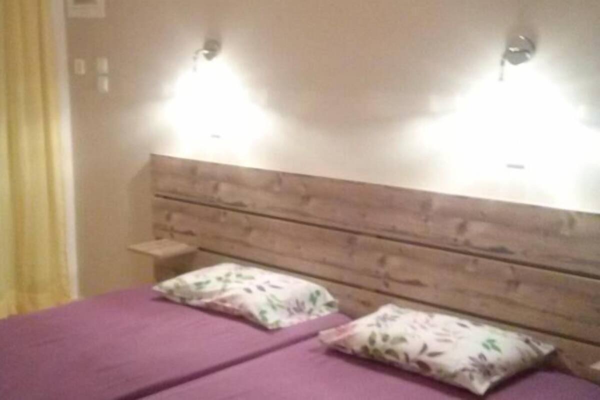 A room with a wooden headboard, two beds with purple bedding, and two wall-mounted lights.