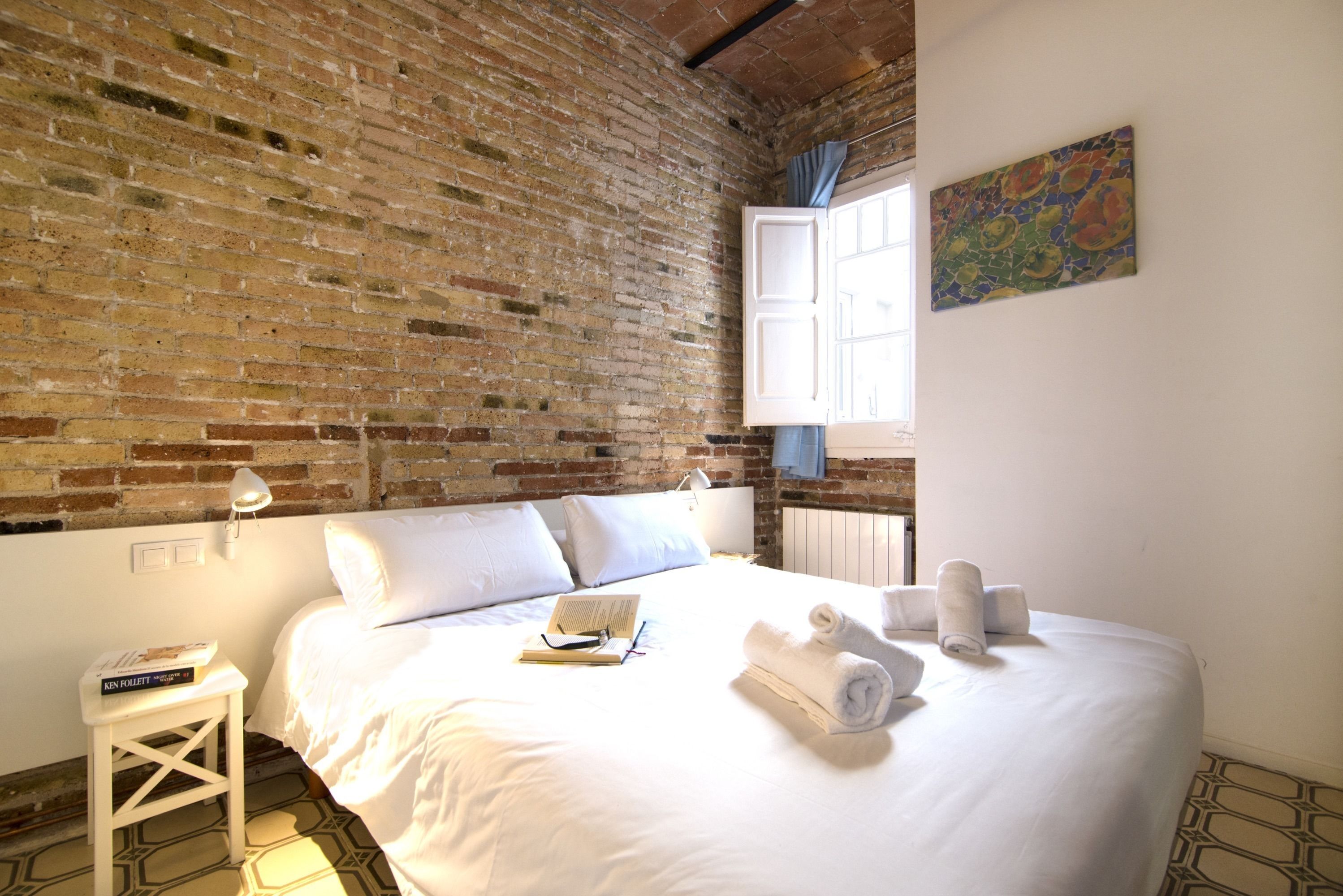 A hotel room with a brick wall, a bed with white linens, a small table with a book, and a painting on the wall.