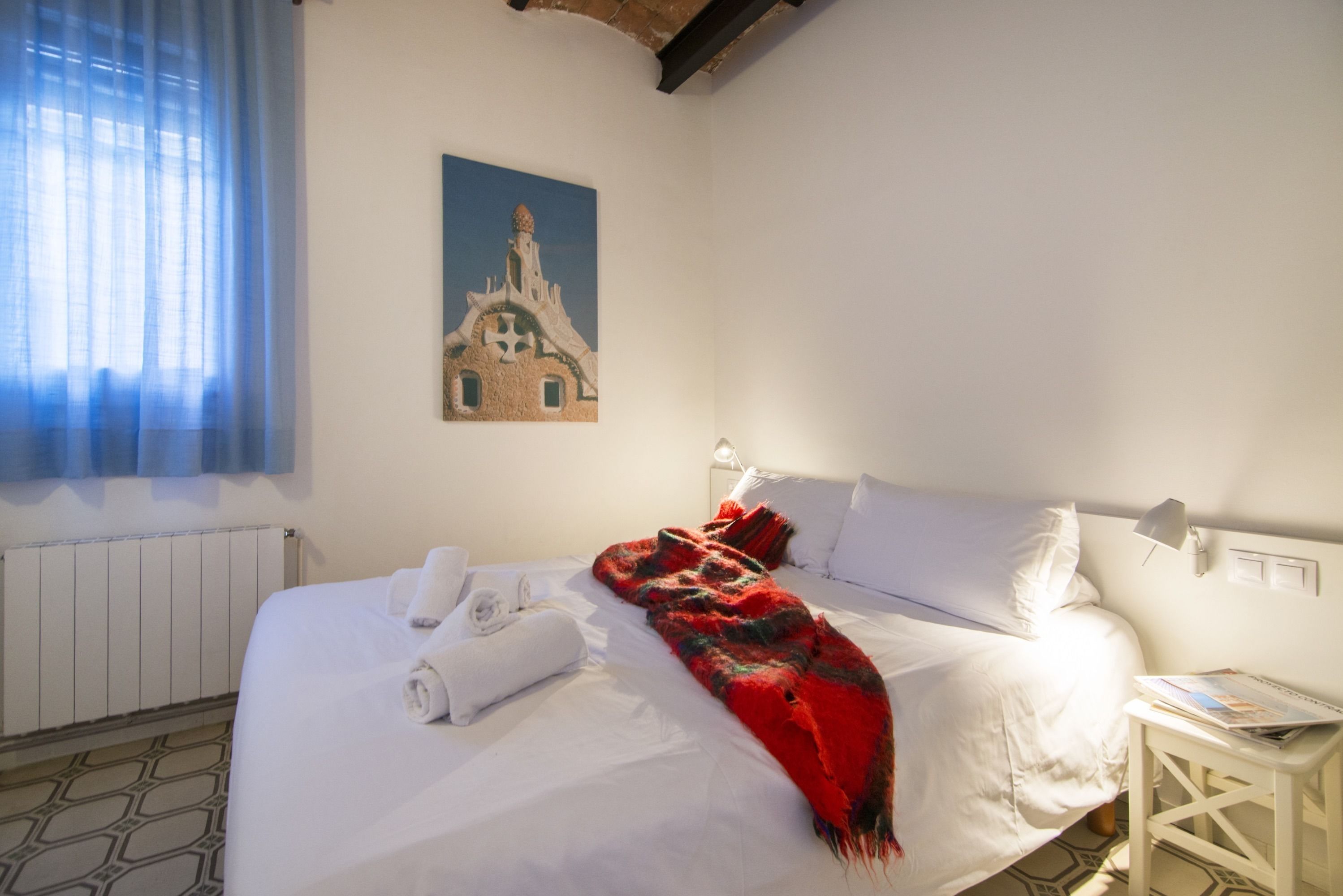 A neatly made bed with white linens and a red blanket, a small bedside table with a lamp, and a painting of a building on the wall.